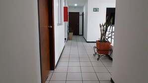 Um corredor com piso de azulejos, porta de madeira, planta em vaso e placa indicando uma saída.