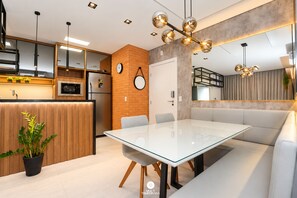 Et moderne køkken med indbyggede hårde hvidevarer, et spiseområde med et glasbord og siddepladser, samt en opholdsstue med sofa og et stort spejl.