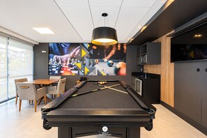 A billiards table with a pool cue, a television screen displaying a game, and a seating area with chairs. - Apto 100m da Praia Com Piscina Aquecida e Lazer - Cachoeira do Bom Jesus - Floripa (Florianópolis)