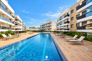 A swimming pool with lounge chairs and umbrellas, surrounded by modern apartment buildings. - Apto 100m da Praia Com Piscina Aquecida e Lazer - Cachoeira do Bom Jesus - Floripa (Florianópolis)