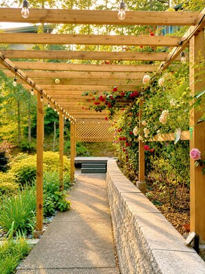 Une pergola en bois décorée de guirlandes lumineuses, un chemin de pierres et des plantes en fleurs.
