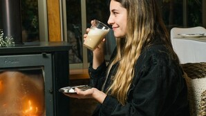 Una persona sentada junto a una estufa de leña, sosteniendo un vaso de bebida.