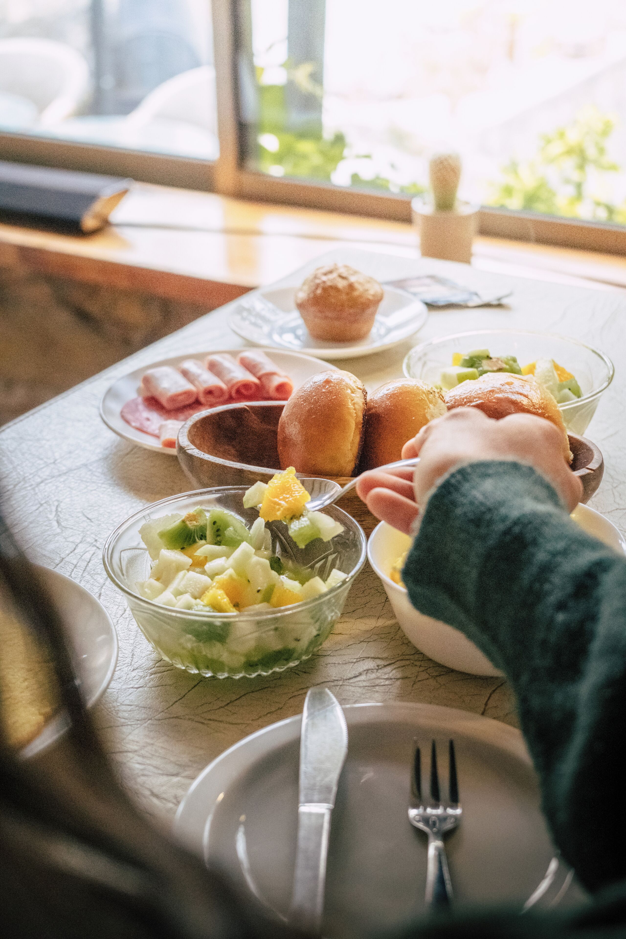 Desayuno con diferentes alimentos sobre una mesa, incluyendo un tazón de ensalada de frutas, panecillos y un muffin.