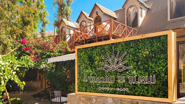 Un muro de piedra con un letrero que dice "El Tesoro de Elqui Hotel-CAFE" y una zona de estar con cojines y un banco tejido.