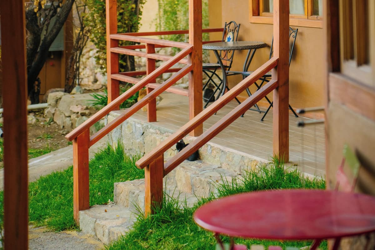 Una escalera de madera conduce a un porche cubierto con una mesa y sillas, rodeado de follaje y un muro de piedra.