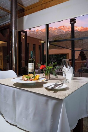Mesa lista para comer con una botella de vino, una copa y un plato de comida, con vista a la montaña al fondo.