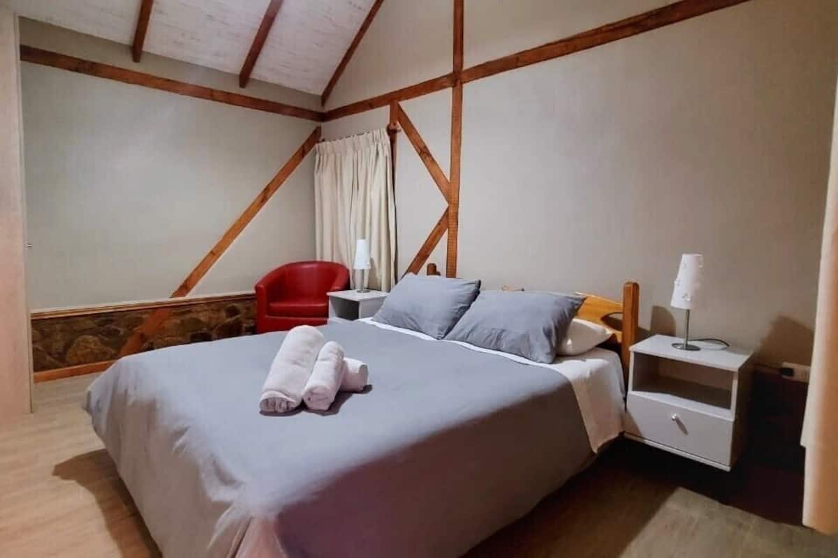 Un dormitorio con cama, mesita de noche, una silla roja y una pared de piedra.