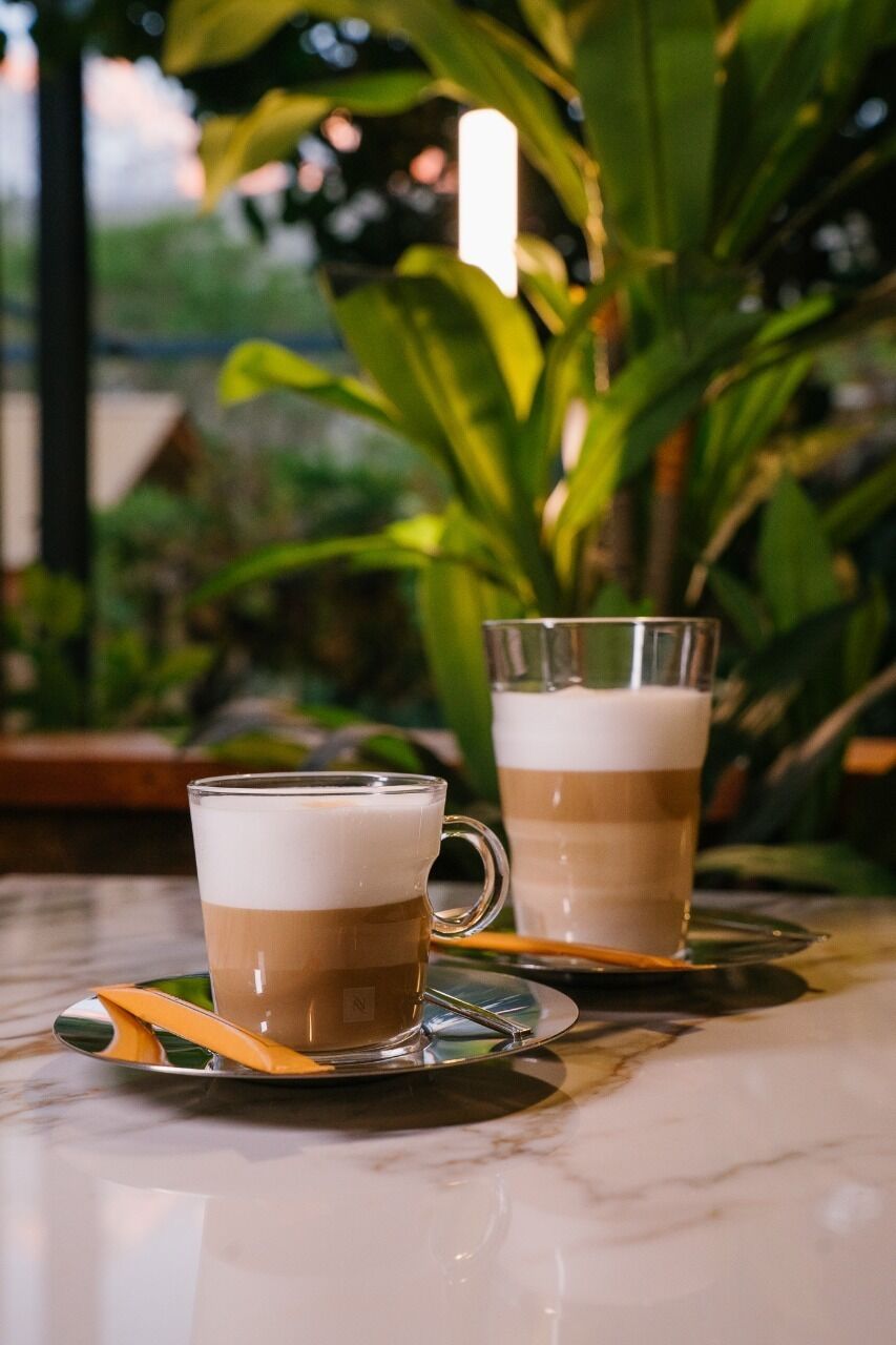Two cups of coffee on a marble table with a spoon and saucer, surrounded by greenery.