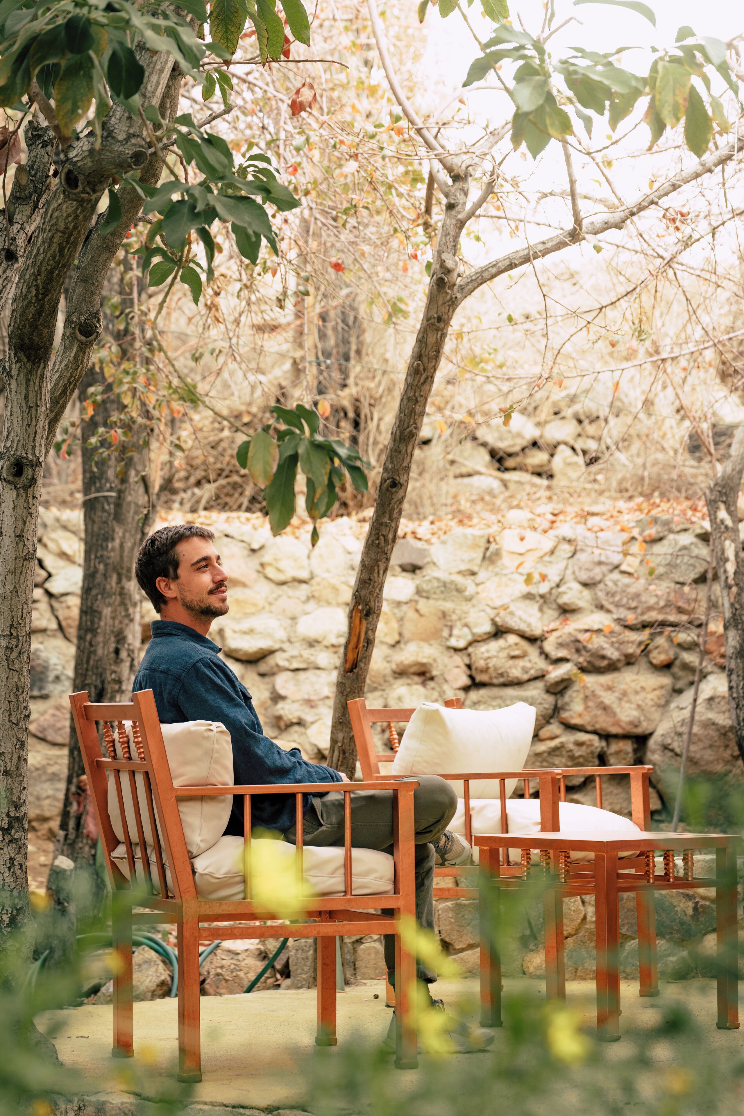A person seated on a wooden bench with a cushion, surrounded by trees and a stone wall.