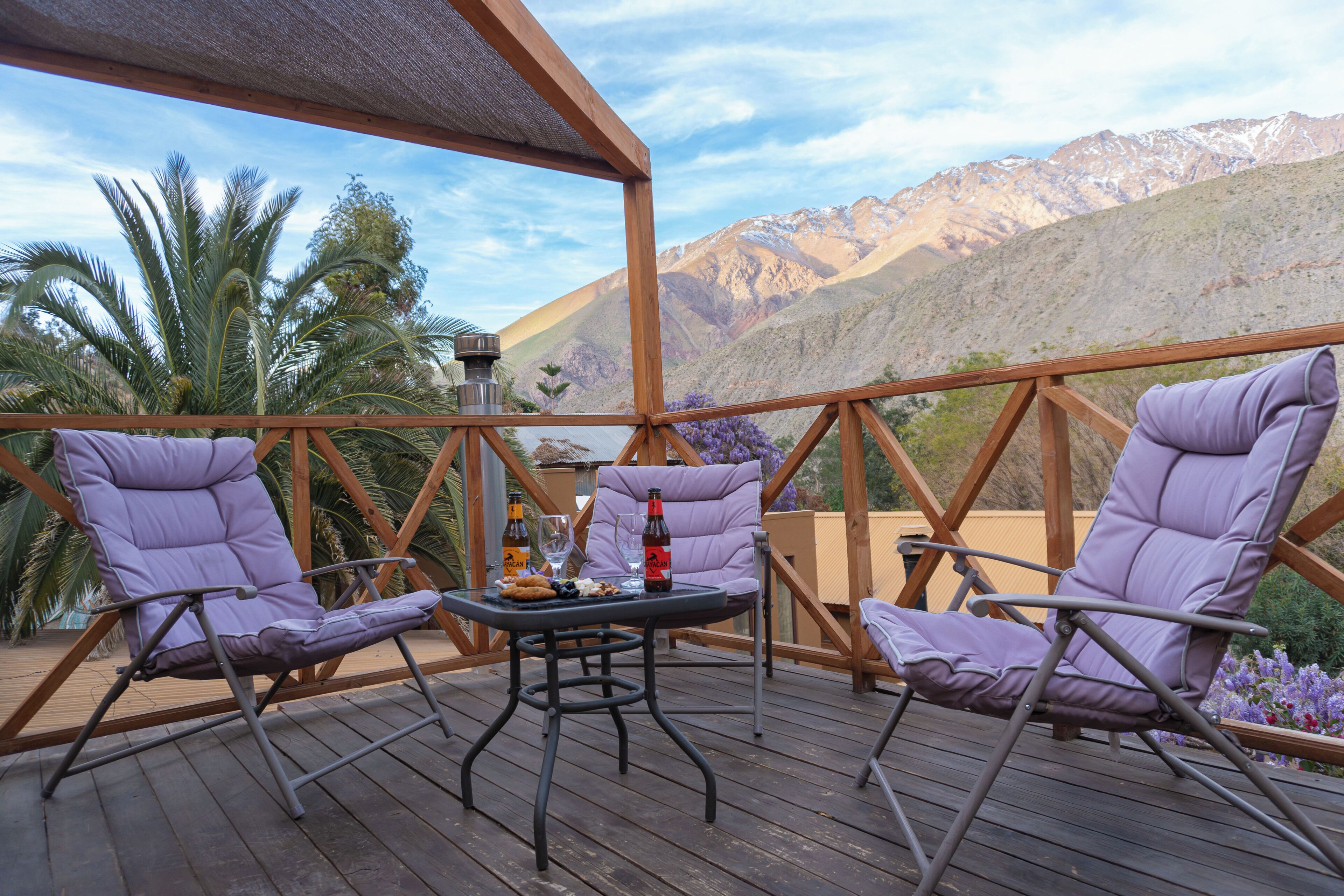 Une terrasse en bois avec deux fauteuils de jardin violets et une table sur laquelle sont disposés des boissons et des en-cas.