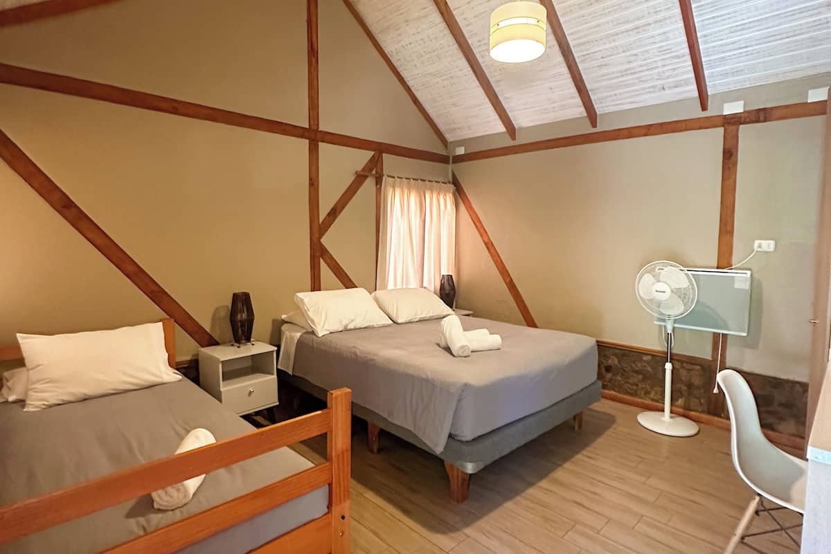 Una habitación con techo inclinado, vigas de madera, dos camas, un escritorio, una silla y un ventilador de pie.