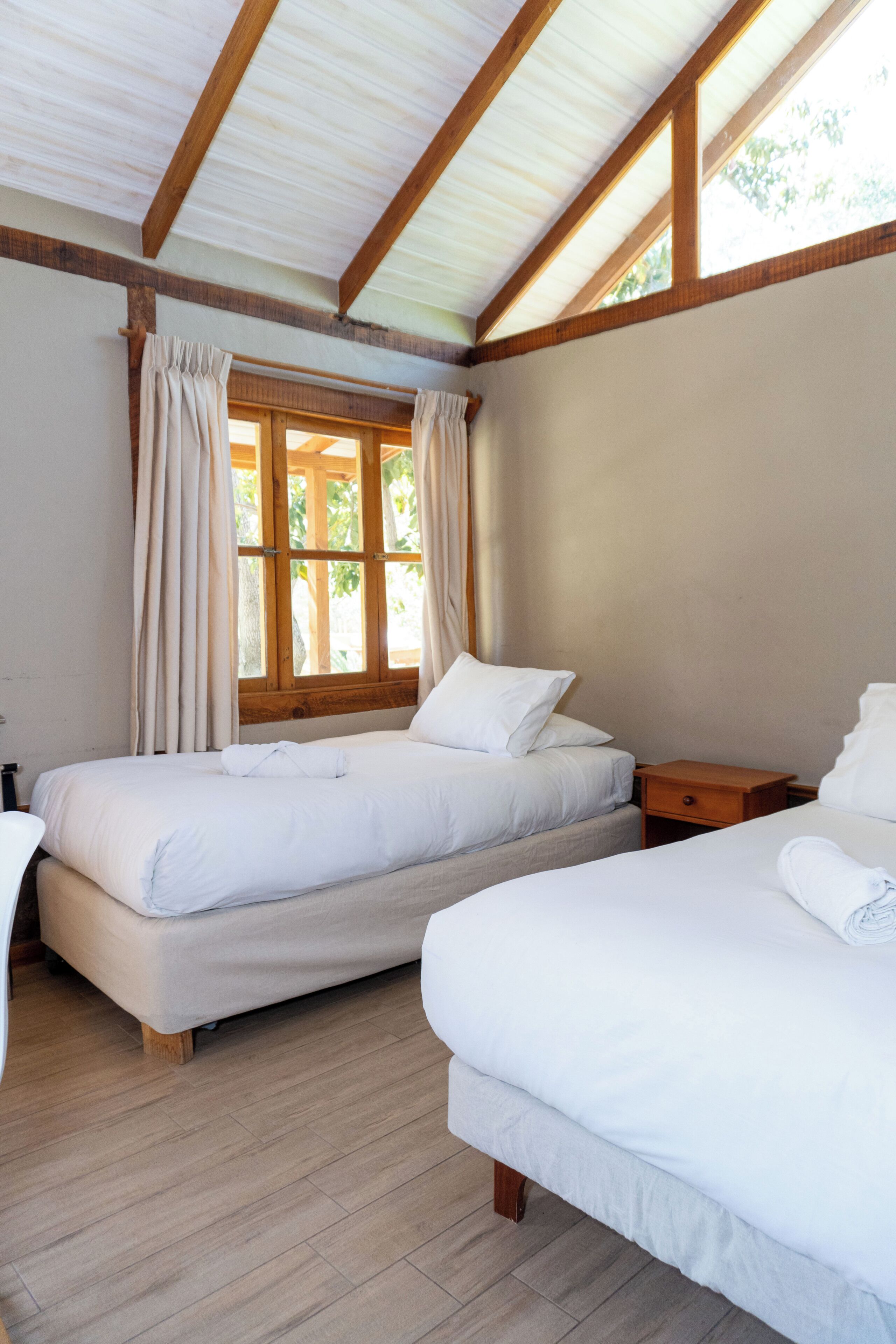 A bedroom with two beds, wooden beams, a window with curtains, and a wooden floor.