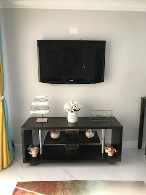 A black entertainment center with a flat-screen TV mounted above it, a small white vase with flowers, and a welcome sign. - The Havens Hideaway (Freeport)