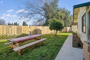 A backyard with a wooden picnic table, a small tree, and a concrete path leading to a building. - Sweet & Cozy, Bird Themed Nest, w/ Hot Tub & Fire Pit. 9 min to Turtle Bay (Redding)