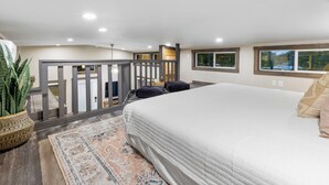 Une chambre avec un grand lit, un plancher en bois, un tapis, une plante en pot et un balcon avec une balustrade.