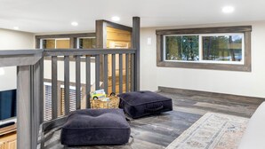 Un intérieur moderne avec un escalier en bois, une grande fenêtre donnant sur la verdure et deux poufs noirs posés sur un tapis à motifs.