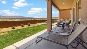 Eine überdachte Terrasse mit Liegestühlen und einem Tisch, mit Blick auf einen angelegten Garten und ferne Hügel.