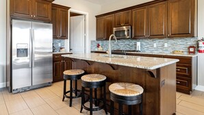 Eine Küche mit Holzausstattung, einem Edelstahlkühlschrank, einer Mikrowelle und einer Arbeitsplatte aus Granit. An der Kücheninsel stehen drei Hocker.