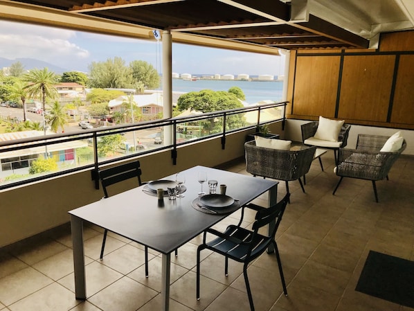 A balcony with a table set for dining, wicker chairs, and a view of the sea and buildings.