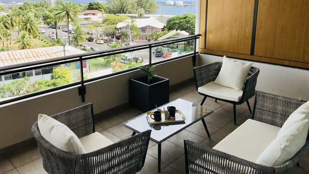 A balcony with wicker furniture, a small table with a tray, and a view of a cityscape with buildings and mountains. - Tipanie- Apt F2 PPT w/pool, A/C, high speed Wifi and wonderful view (Papeete)