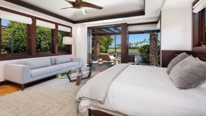 A modern bedroom with a large bed, a sofa, and a glass coffee table. There is a view of a garden and a pool outside. - Unmatched location Four Seasons Hualalai  5BR/5BA Ocean Estate 6500sqft 3/4 acre (Kailua-Kona)