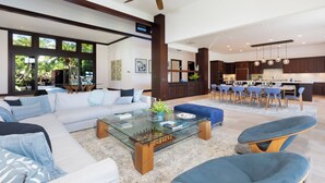 A spacious living area with a large sofa, a glass coffee table, and a dining area with blue chairs. - Unmatched location Four Seasons Hualalai  5BR/5BA Ocean Estate 6500sqft 3/4 acre (Kailua-Kona)