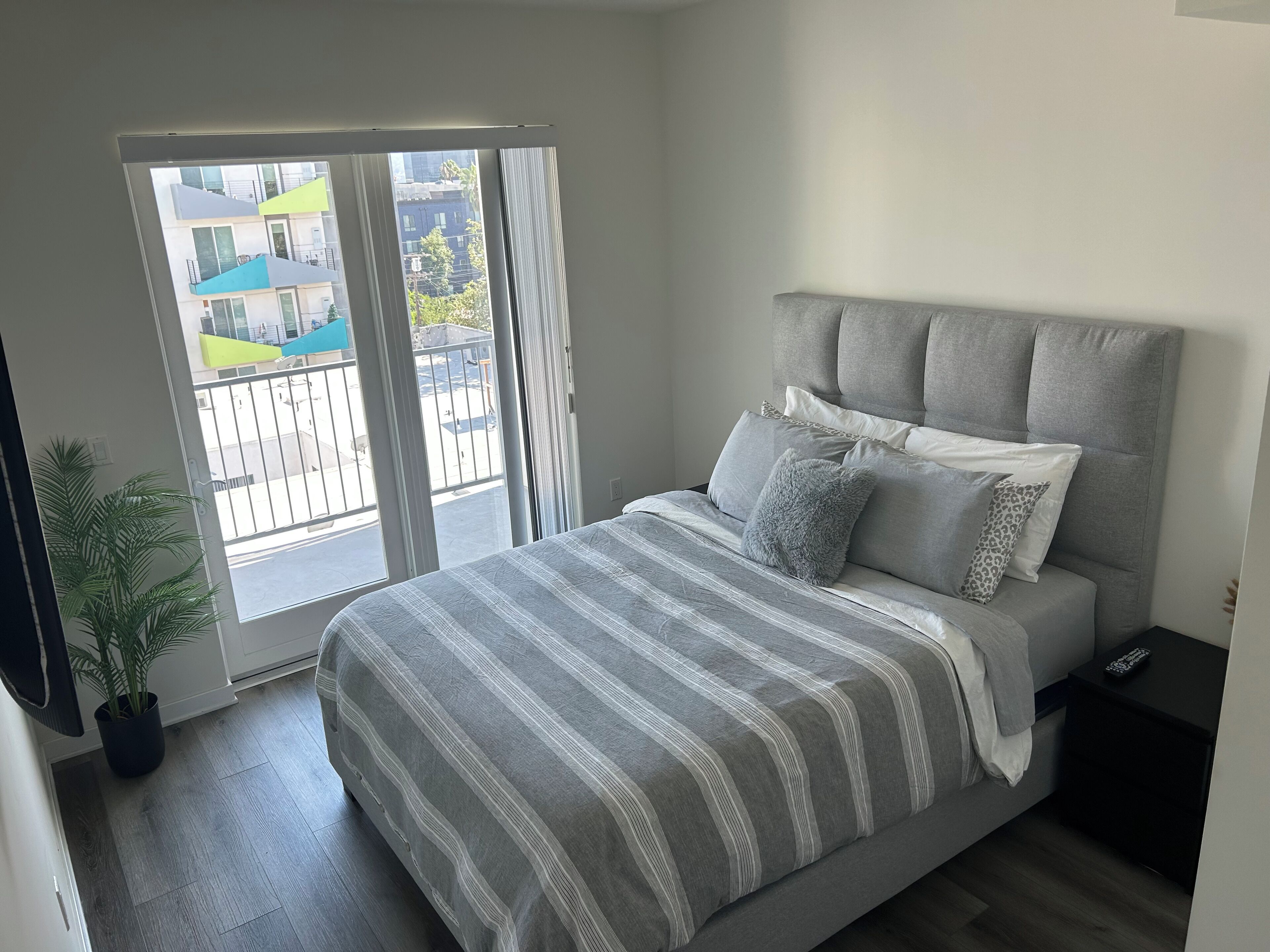 Ein Schlafzimmer mit einem Bett, einem Nachttisch, einer Topfpflanze und einer Glastür, die auf einen Balkon führt.