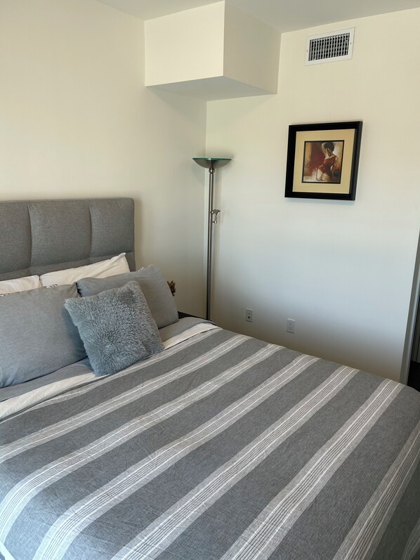 En pænt redt seng med stribet sengetøj, en grå sofa med puder, en gulvlampe og et indrammet billede på væggen.
