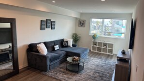 A modern living room with a grey sectional sofa, a small round coffee table, and a large window with a city view. - Spacious & Modern 1BedRM in Noho (Burbank)