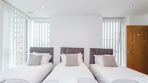 Een hotelkamer met drie bedden, elk voorzien van een grijs hoofdbord, wit bedlinnen en een raam met verticale jaloezieën.