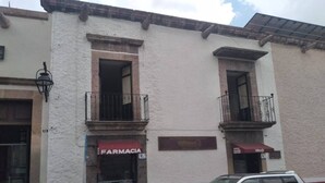 Un immeuble avec une pharmacie au rez-de-chaussée, une enseigne indiquant « ESPECULATAS » au-dessus de l’entrée, et un VUS argenté garé devant.