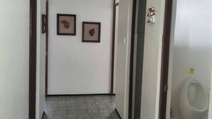 Un couloir au sol carrelé, deux tableaux encadrés accrochés au mur et un urinoir dans la pièce adjacente.