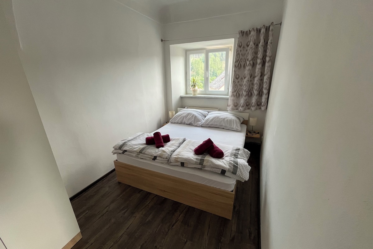 A single bed with a wooden frame, white bedding, and two red towels. A window with a view of greenery is above the bed.