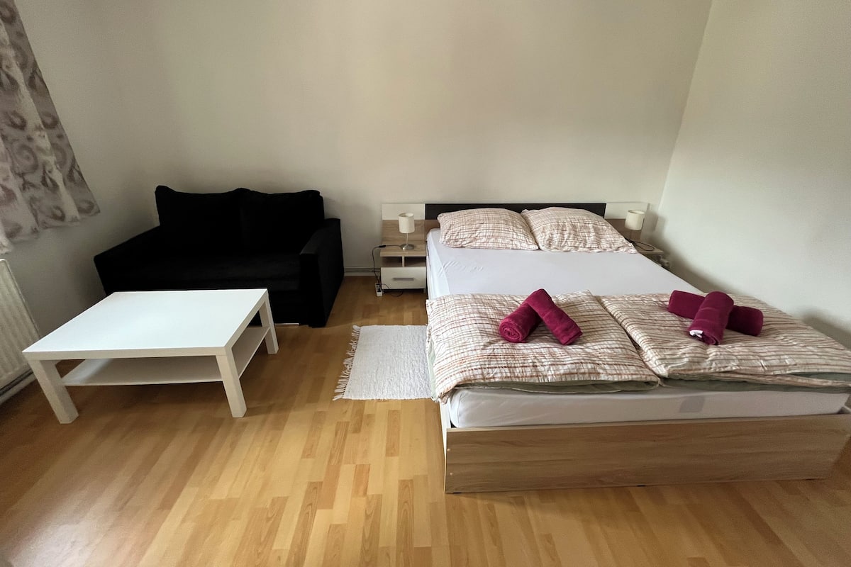 A room with a bed, a black sofa, a small white table, and a bedside table with a lamp.