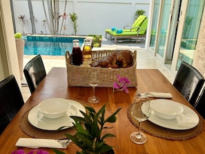 A dining table set with plates, glasses, and a basket of pastries, overlooking a pool and lounge area.