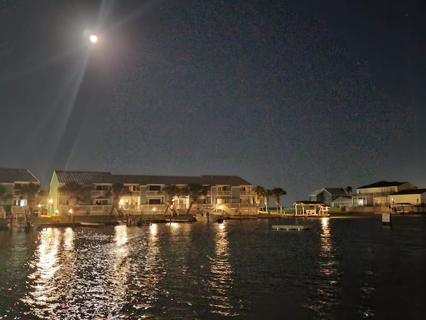 Ein Hotel am Wasserufer mit mehreren Gebäuden, spiegelnder Wasseroberfläche und sichtbarem Mond am Himmel.