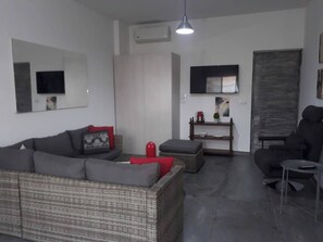 Uma sala de estar com um sofá, um banquinho vermelho, uma mesa pequena com uma chaleira e uma televisão presa à parede.