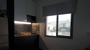 A small, dimly lit room with a bed, a kitchenette with a tiled backsplash, and a window overlooking a cityscape. - Studio In Dbayeh In A Prime Location, Wifi, 38sqm (Dbayeh)