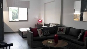 A modern living room with a sofa, a coffee table, a red vase, and a window with blinds. - Studio In Dbayeh In A Prime Location, Wifi, 38sqm (Dbayeh)