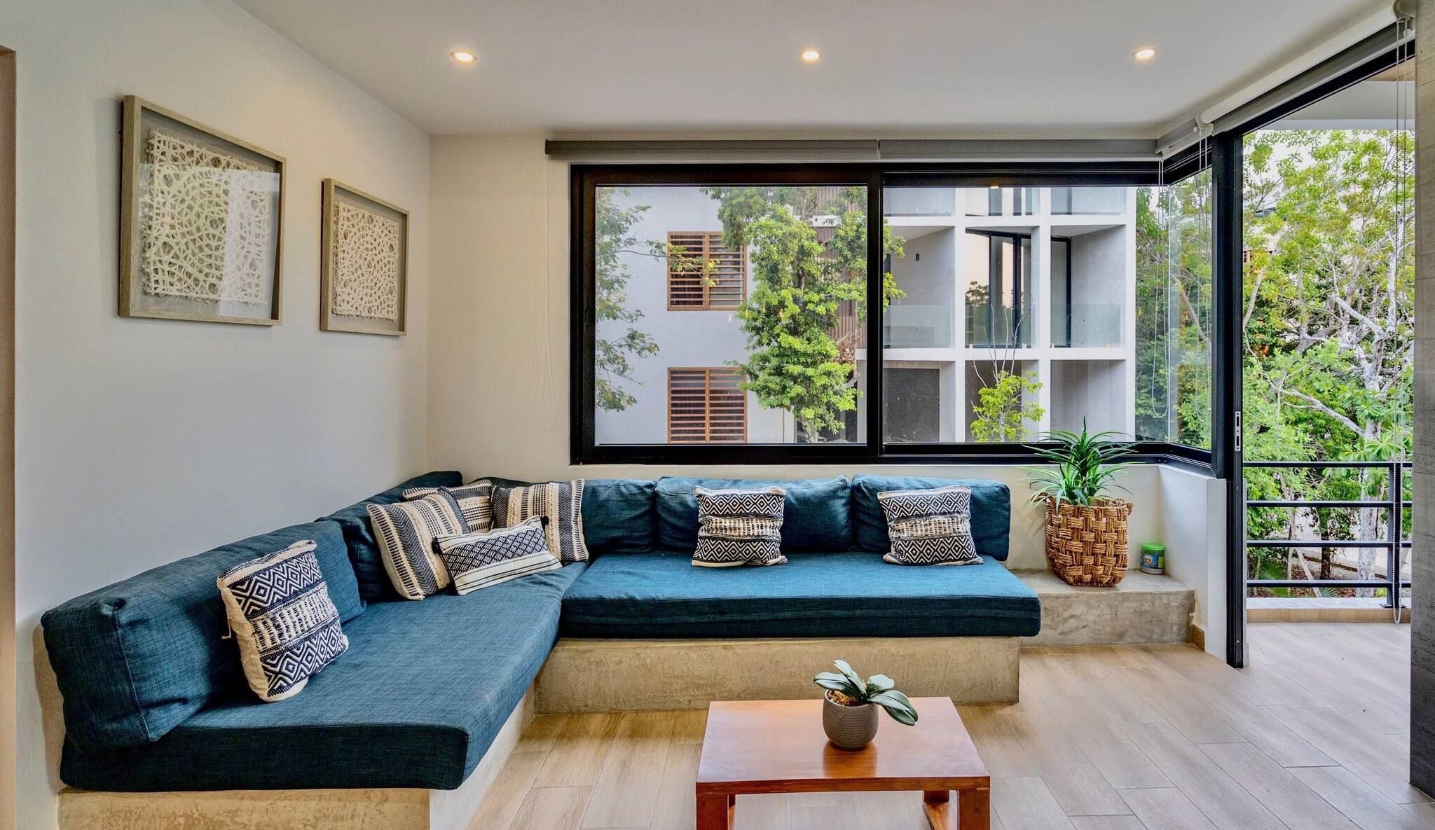 A modern living room with a sectional sofa, a wooden coffee table, and a large window offering a view of buildings and trees.