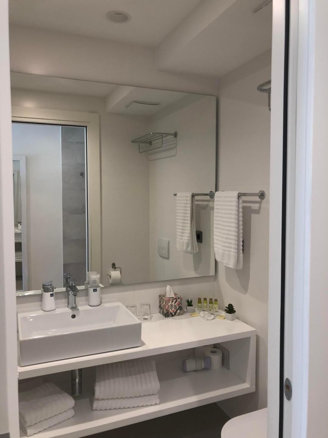 A modern bathroom with a large mirror, two sinks, a towel rack, and a shelf with toiletries.