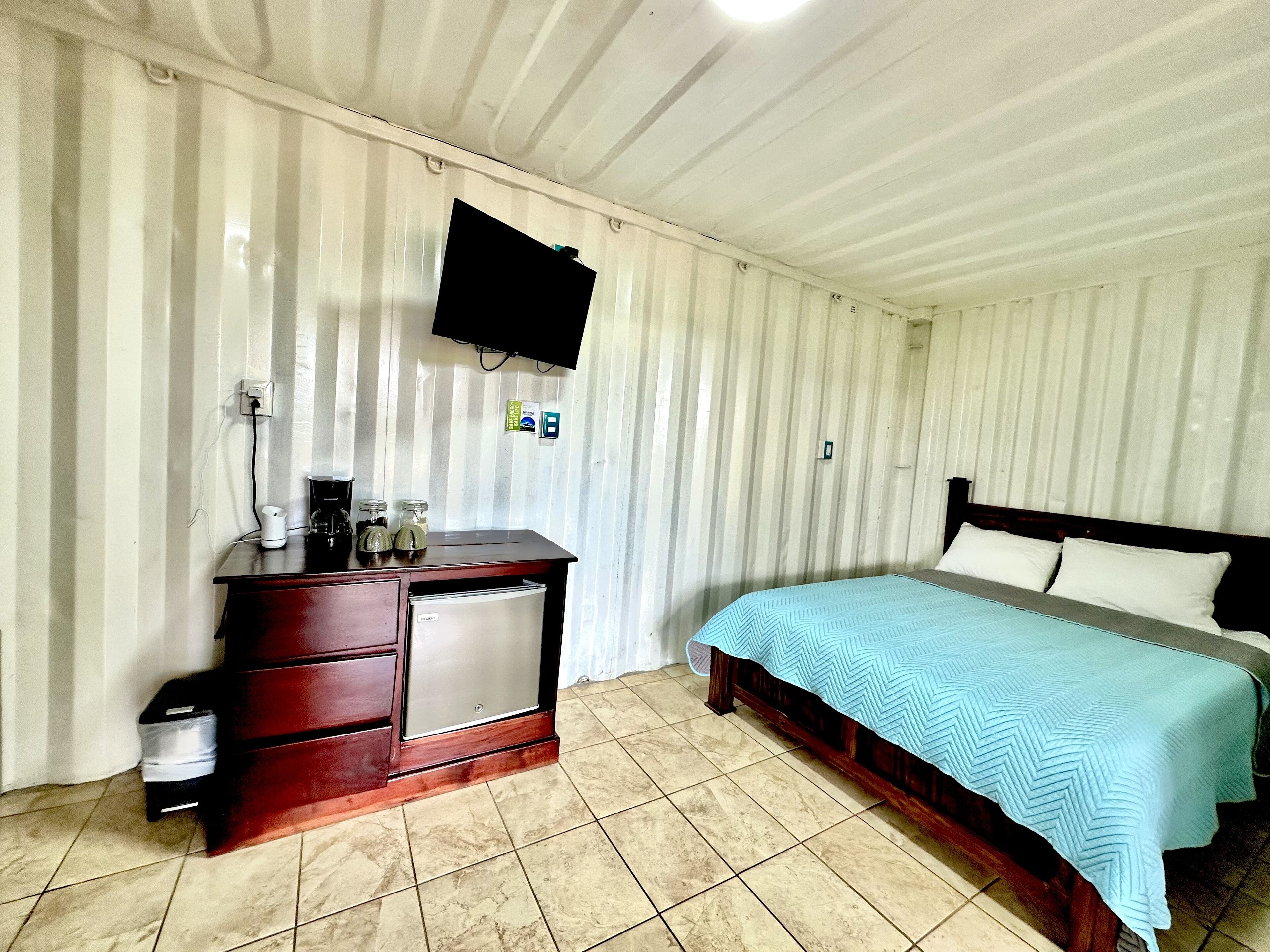 Une chambre à coucher comprenant un lit, une commode en bois, un petit réfrigérateur, un téléviseur fixé au mur et un sol carrelé.