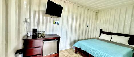 Une chambre à coucher comprenant un lit, une commode en bois, un petit réfrigérateur, un téléviseur fixé au mur et un sol carrelé.