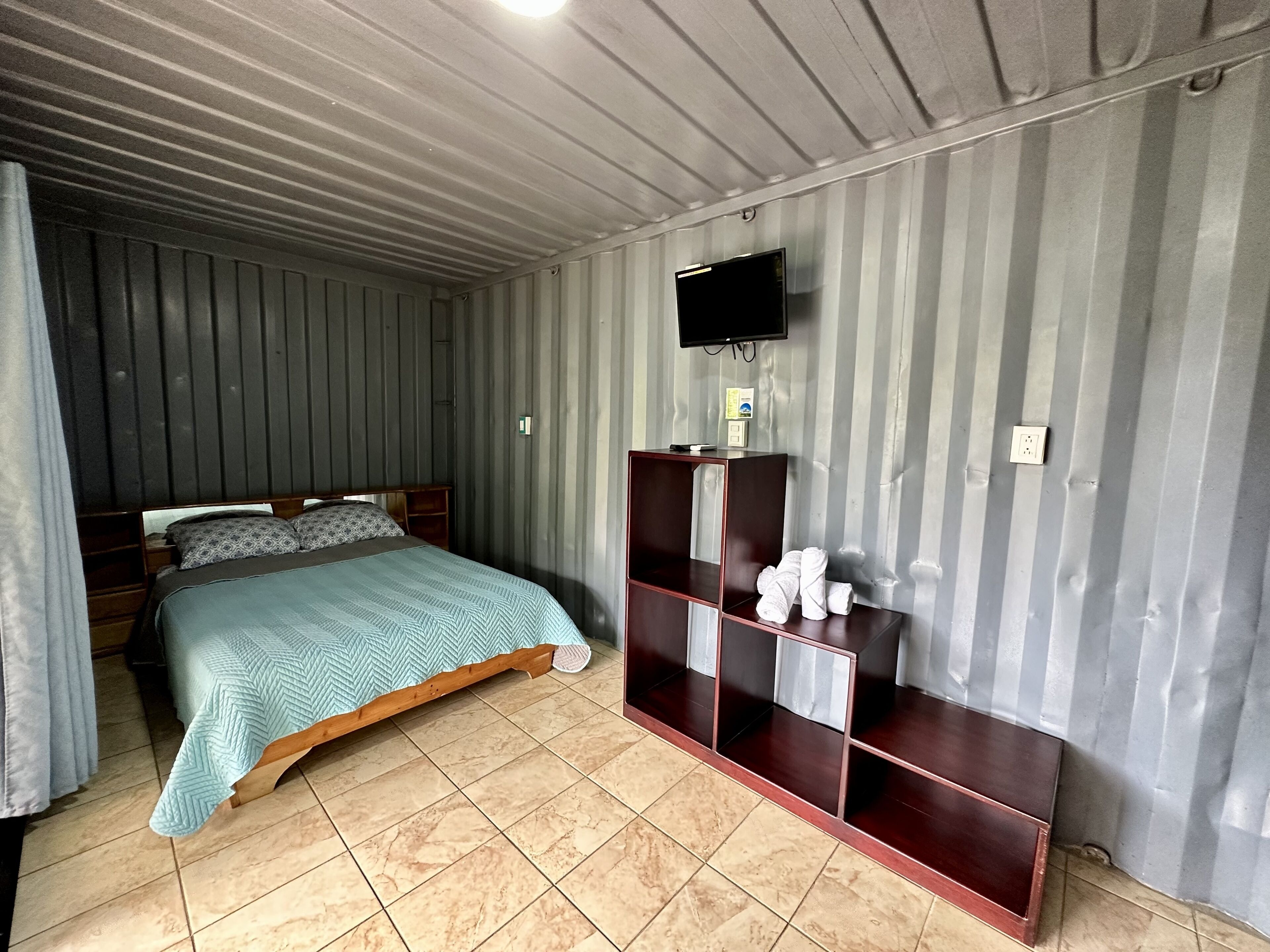 A room with a bed, a TV mounted on the wall, and a wooden cabinet with towels.