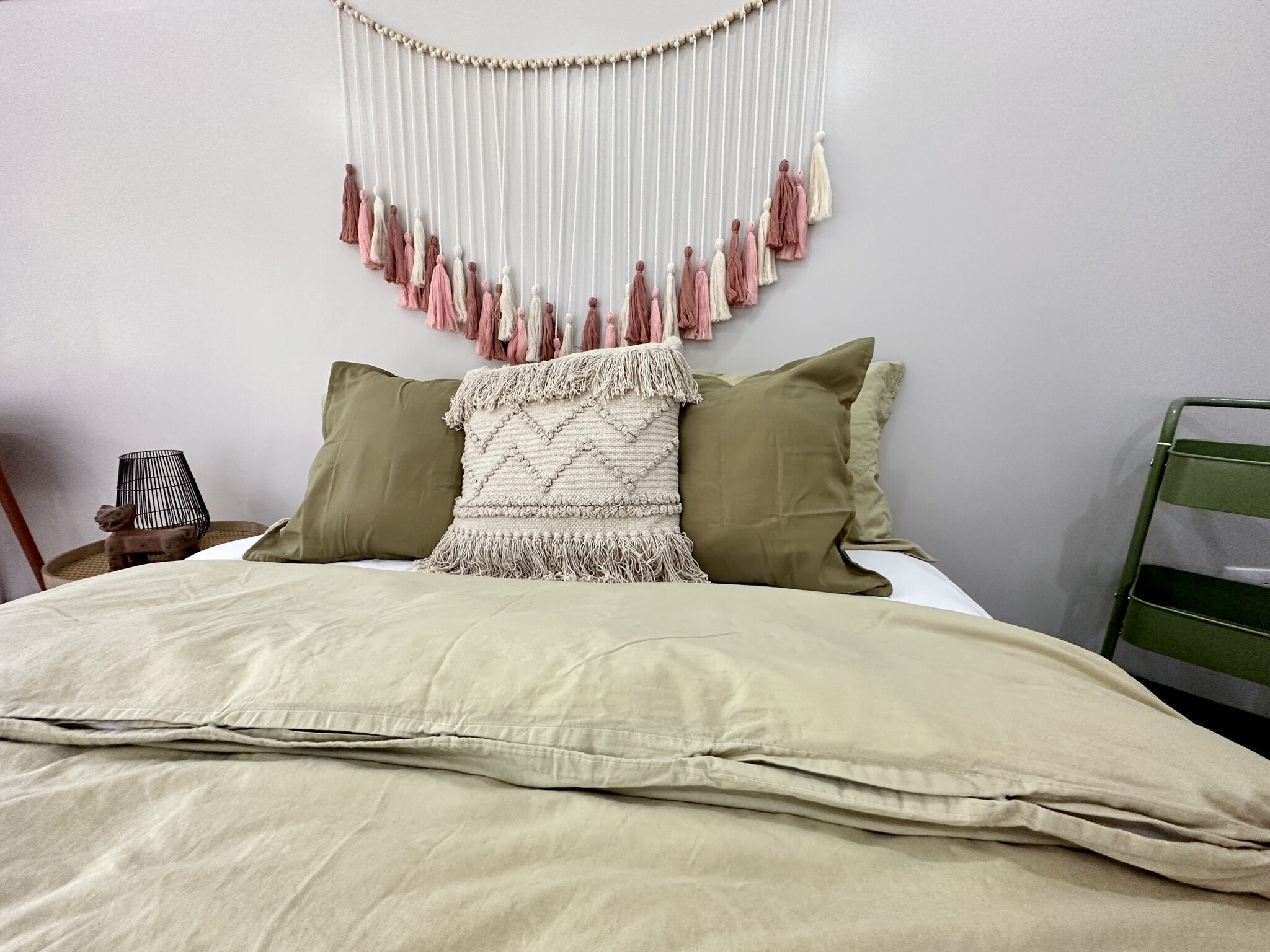 A neatly made bed with a beige blanket, two pillows, and a decorative macramé wall hanging.
