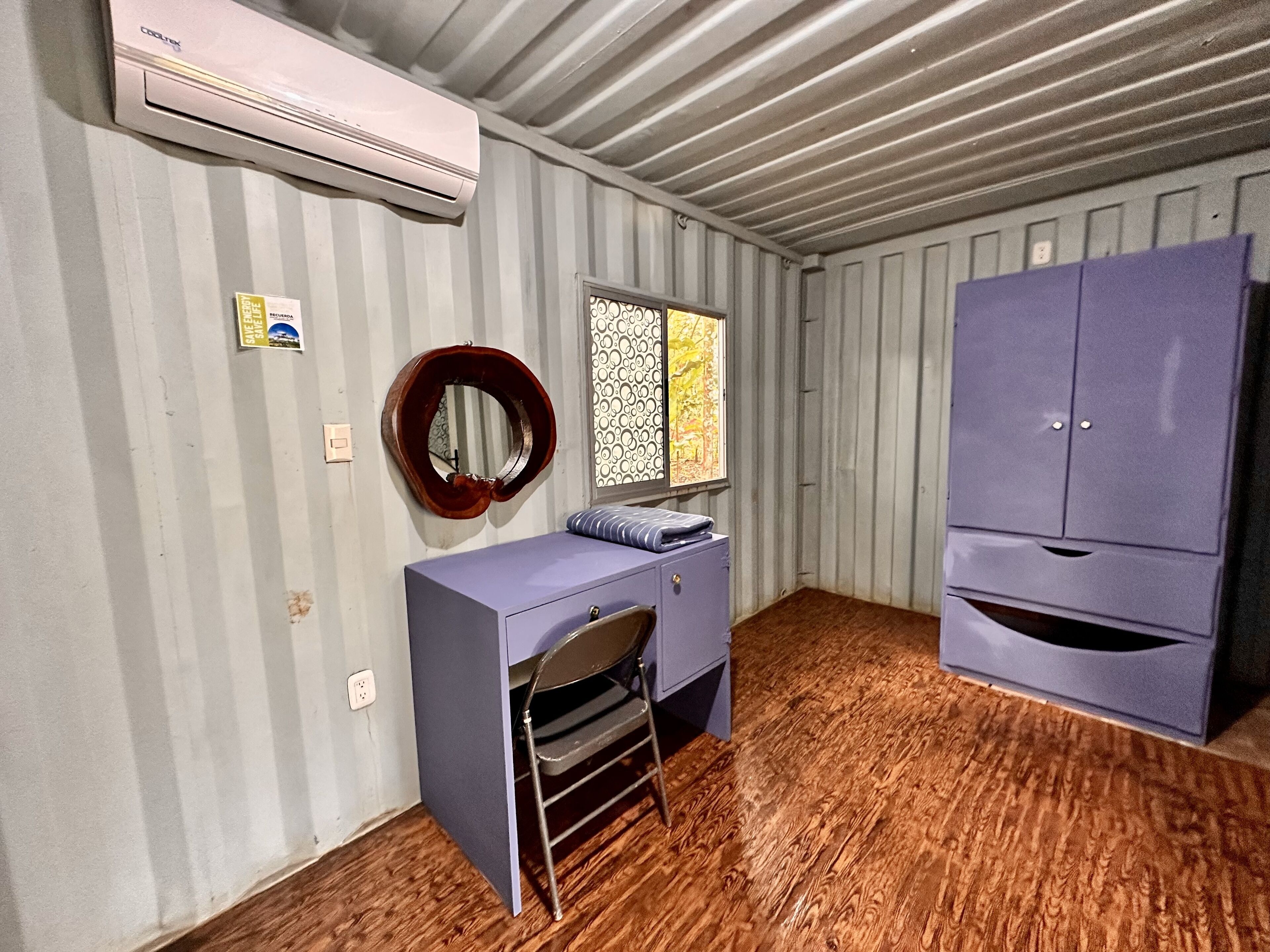 A compact room with a wooden floor, a desk with a chair, a wardrobe, a mirror, and a window with patterned curtains.