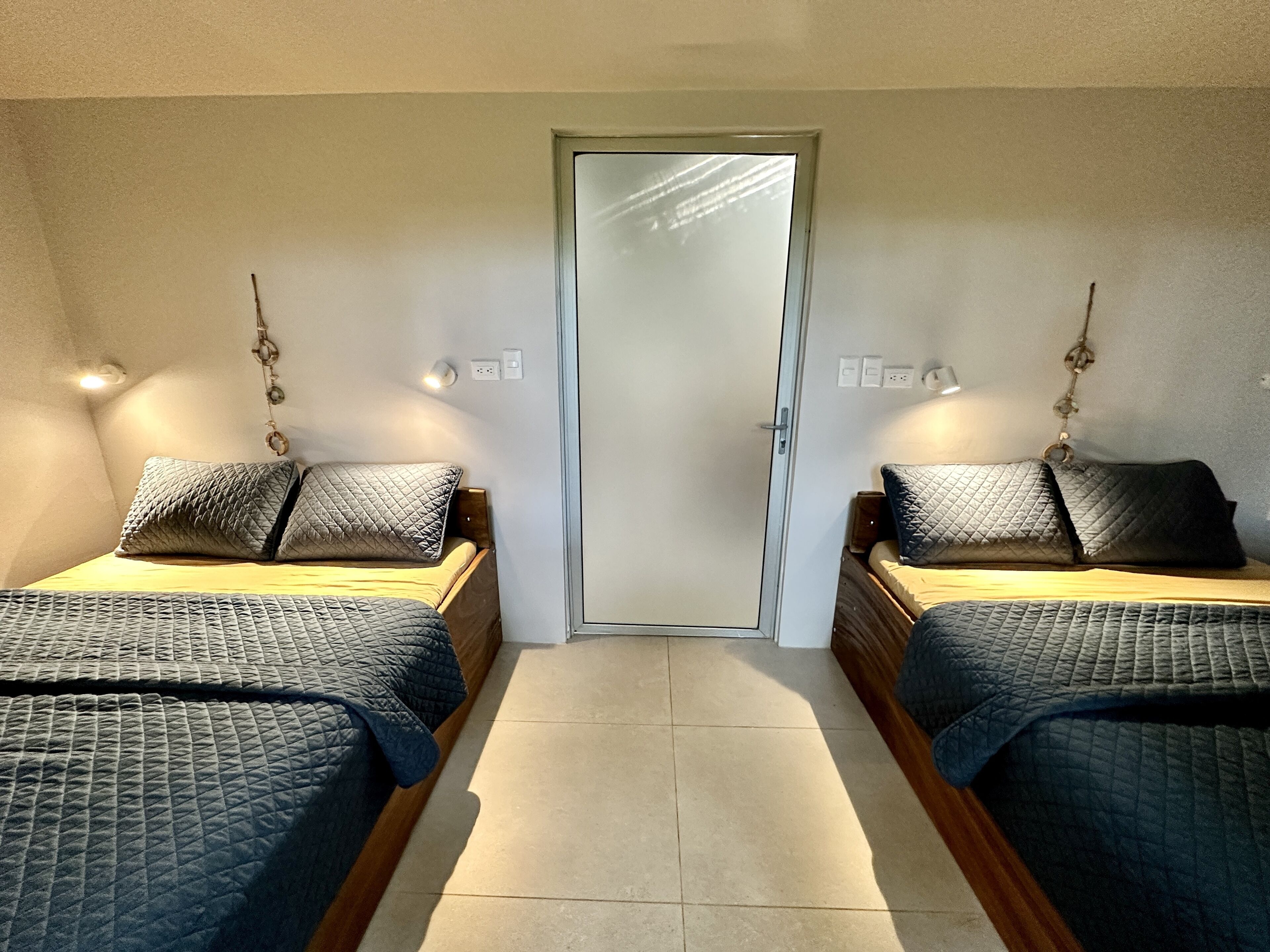 A room with two beds, each with a quilted bedspread and a pillow. There are wall-mounted lights on either side of the door.