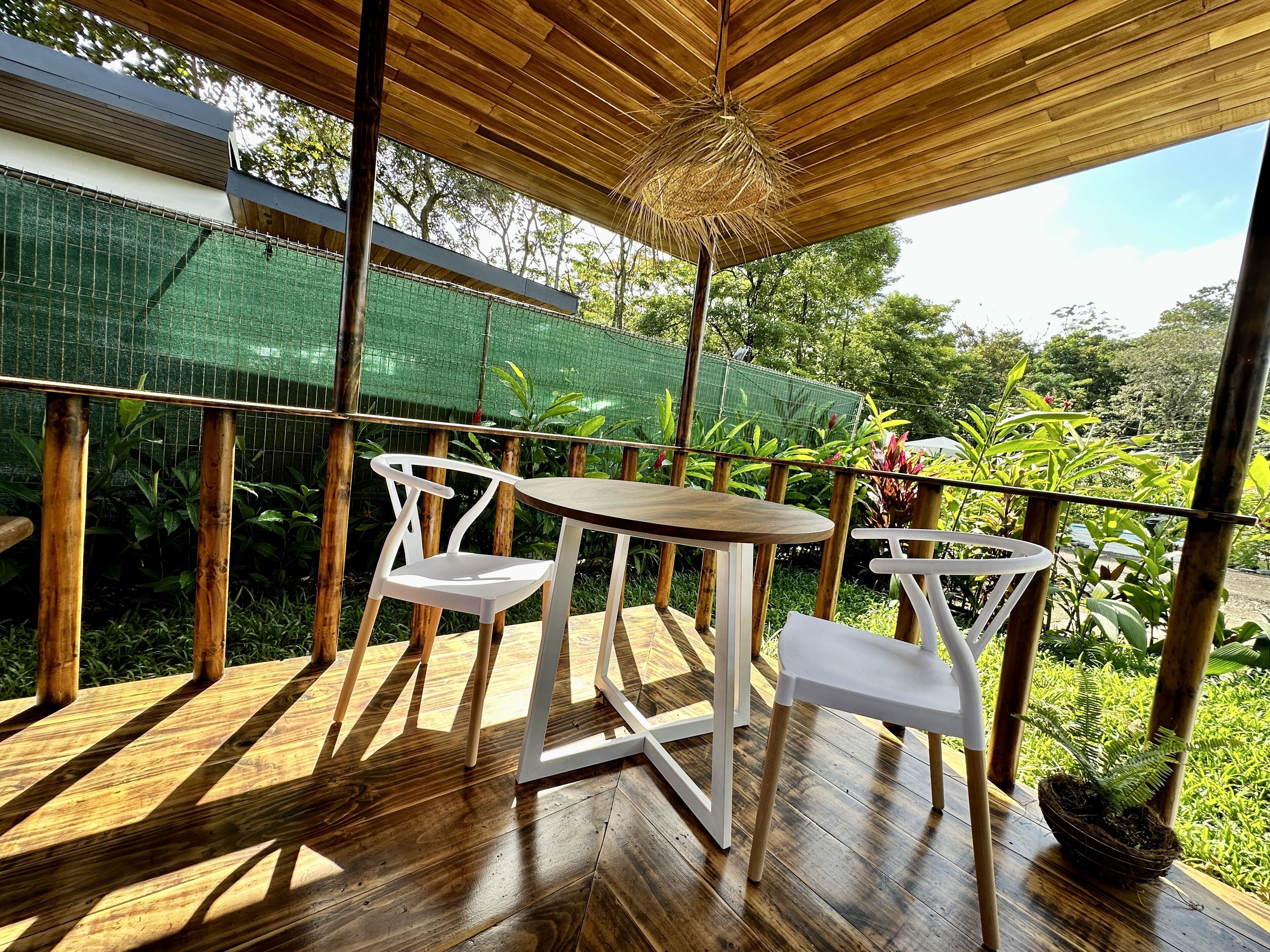 An outdoor wooden deck with a table and two chairs, surrounded by greenery and a green mesh fence.