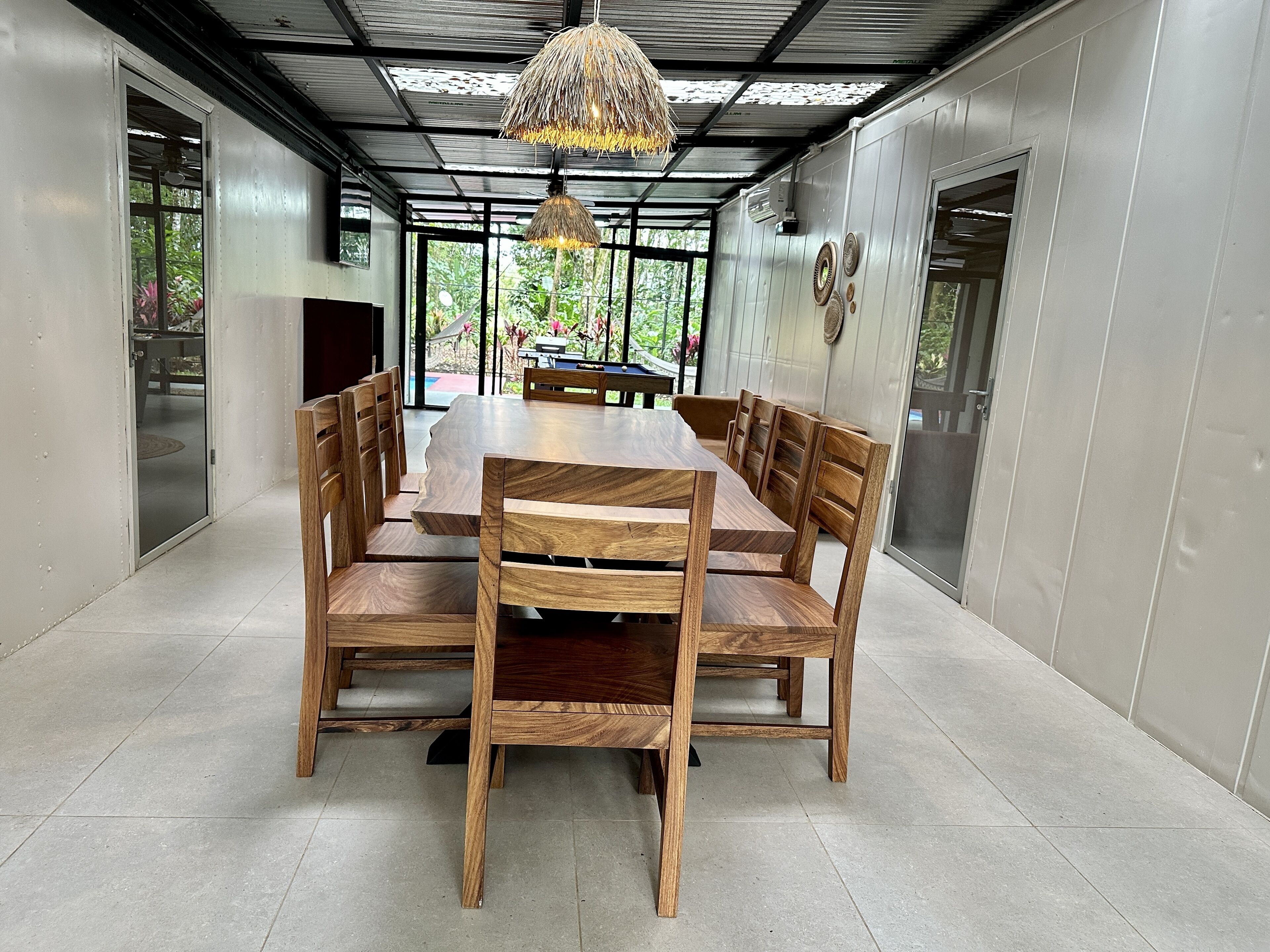Un'area pranzo moderna con un tavolo di legno grande e sedie, un lampadario a sospensione e vista sul verde all'esterno.