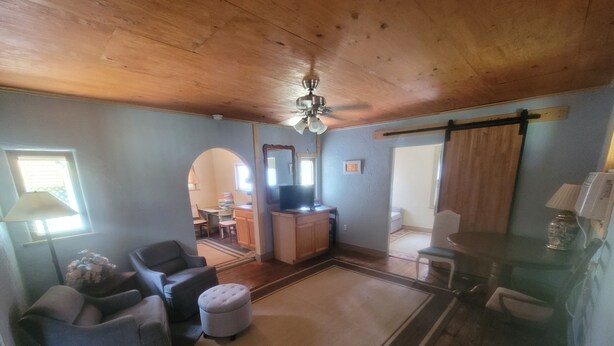 A spacious living room with a wooden ceiling, a ceiling fan, a television, and a dining area visible through an archway.
